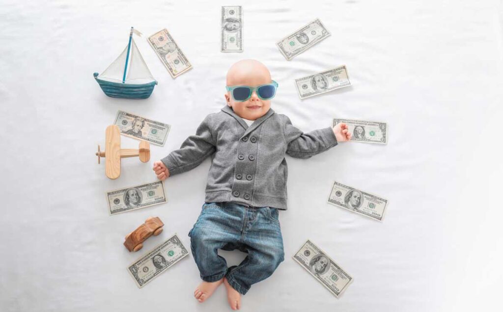 Ein Baby mit Sonnenbrille liegt in einem Kreis aus Geldscheinen, Segelboot, Flugzeug und einen Auto. (c) AdobeStock