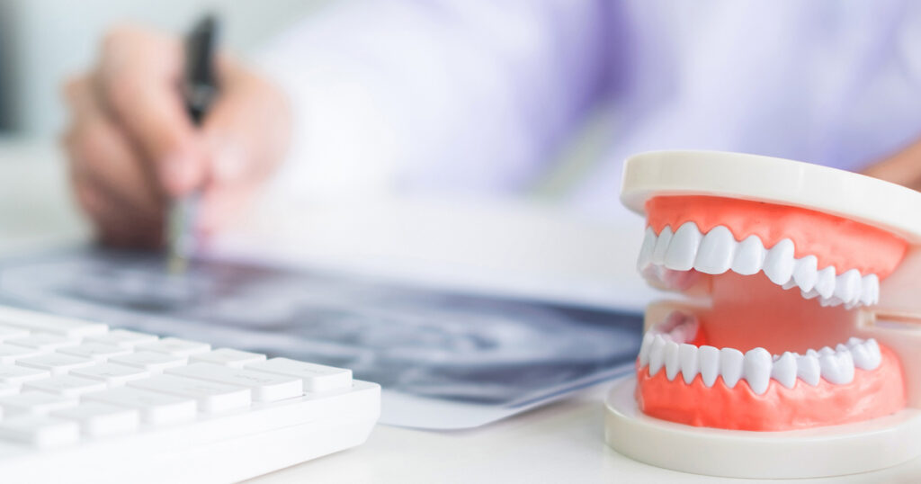 Concentrated dentist sitting at table with jaw samples tooth model in dental office professional dental clinic. (c) AdobeStock