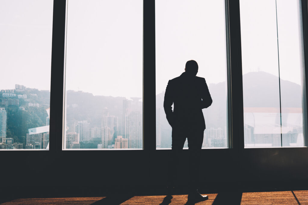 Back view of businessman in elegant suit standing indoors in loft interior.Male entrepreneur dressed in formal wear looking out of window enjoying scenery and thinking on building project in office. (c) AdobeStock
