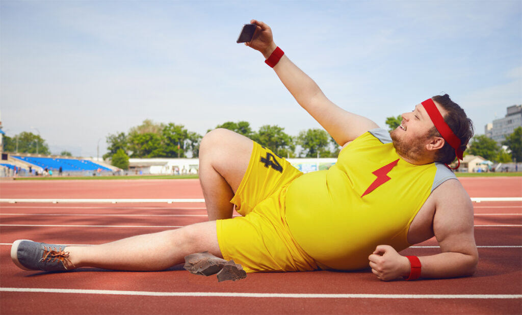 Fat funny man lying down doing a selfie on the phone on the track in the stadium. (c) AdobeStock