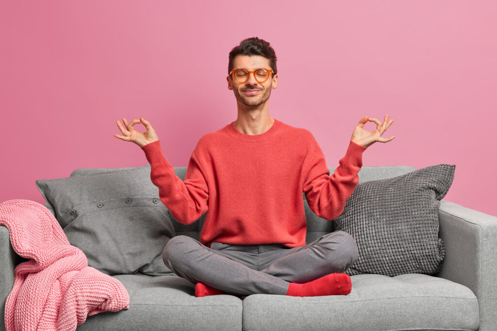 Pleased young European man with closed eyes sits on sofa in lotus pose meditates at home dressed in casual clothes against pink background practices yoga. People meditation wellness concept. (c) AdobeStock