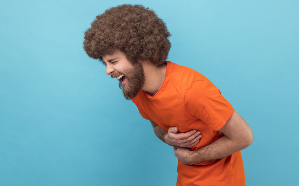 Side view of laughing man with Afro hairstyle in orange T-shirt holding his stomach and hunched in crazy hysterical laughter, sincere joyful emotions. Indoor studio shot isolated on blue background. (c) AdobeStock
