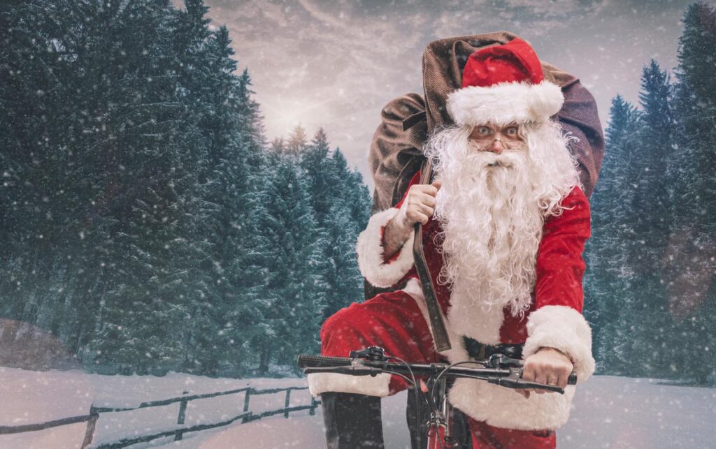 Santa Claus riding a bicycle and carrying a heavy sack with gifts for Christmas under the snow. (c) AdobeStock