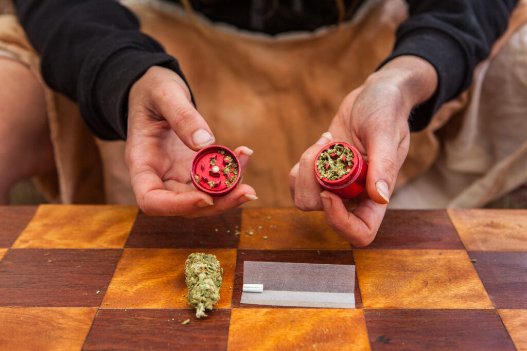 Selective focus closeup of person's hands holding a red weed filled grinder shot from above. Checkered table with rolling paper, filter and bud displayed below. (c) AdobeStock