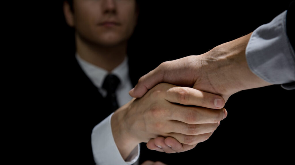 Anonymous business partners making handshake in dark shadow. (c) AdobeStock