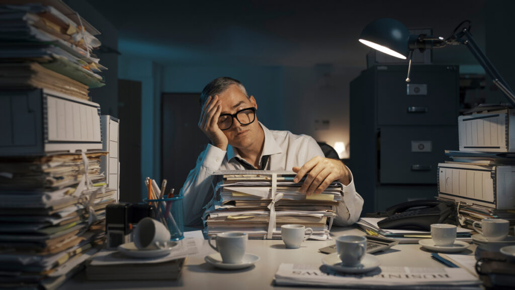 Exhausted businessman sleeping at his desk and working overtime late at night, he is surrounded by piles of paperwork. (c) AdobeStock