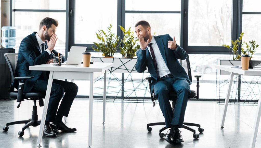 Zwei Männer in einem stylischen Büro beim Tratschen über einen Schreibtisch hinweg. (c) AdobeStock