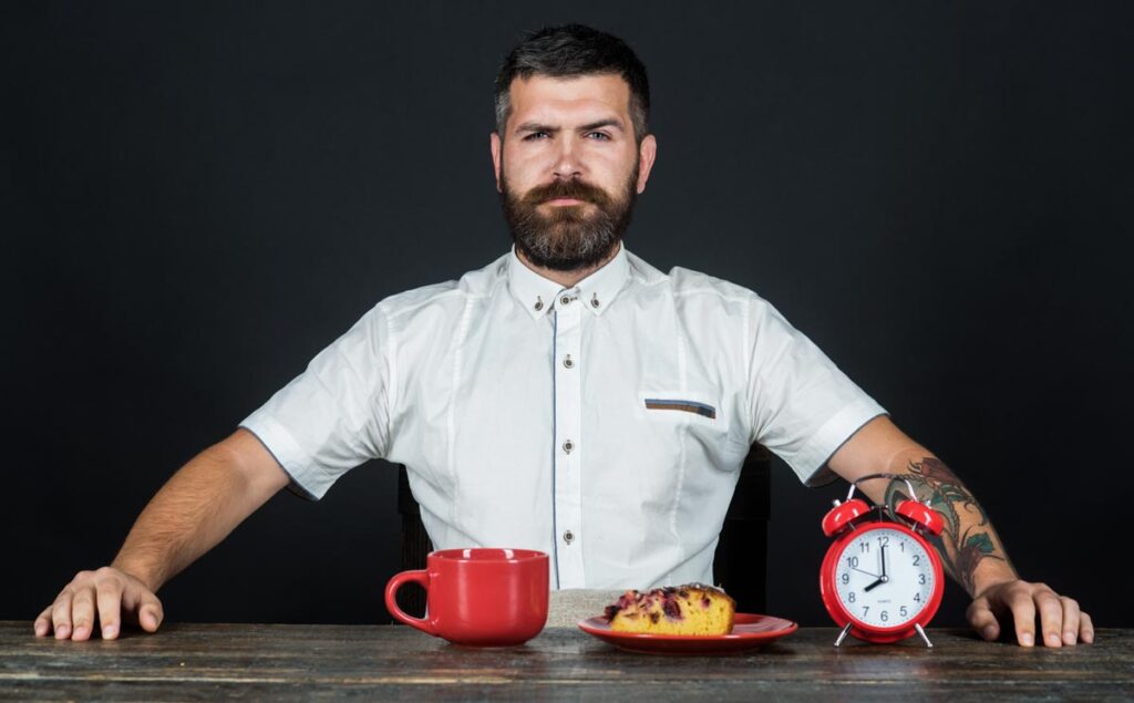 Ein Mann mit weißem Hemd und Vollbart an einem Tisch mit einer großen Tasse, einem Stück Kuchen und einem Wecker. (c) AdobeStock