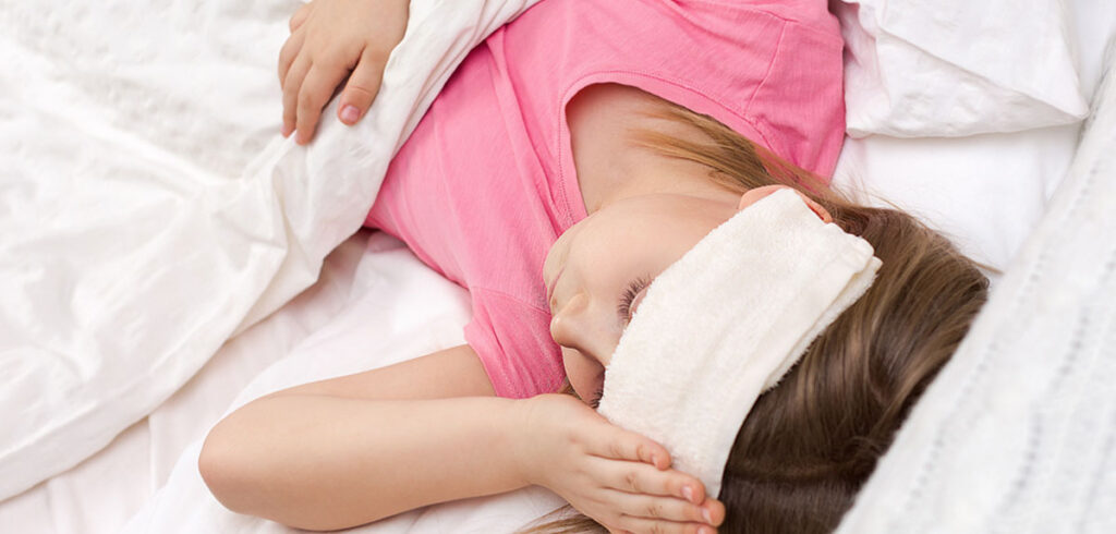 Ein Kind mit einem Tuch auf der Stirn in einem Bett. (c) AdobeStock