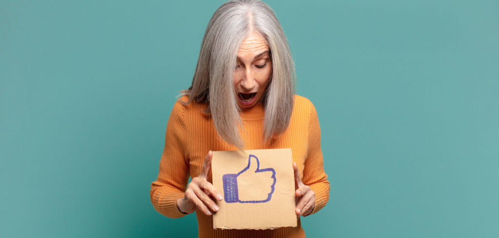 DIE »goldene« Social-Media-Regel Nr. 1: Halte deine Mutter von Facebook fern Eine Frau mit langen grauen Haaren hält ein Schild mit einem Facebook-Daumen hoch. (c) AdobeStock