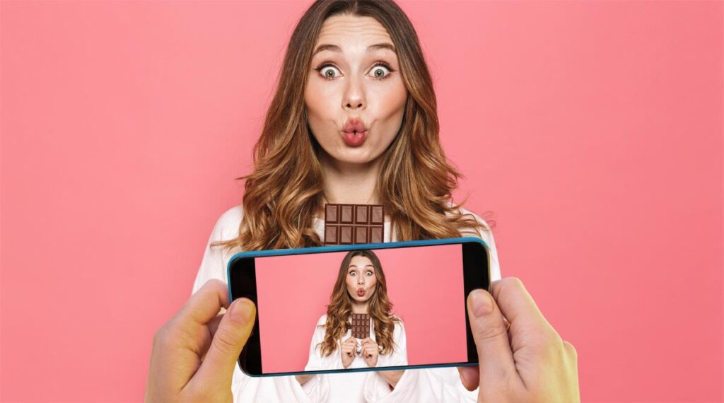 Eine junge Frau mit einer Tafel Schokolade, die gerade mit einem Handy fotografiert wird. (c) AdobeStock/ K2