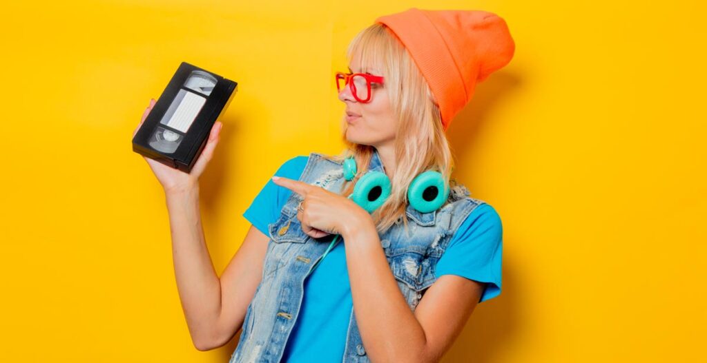 Anleitung zum »wirklich« Coolsein 1 Eine Frau mit roter Brille, oranger Mütze und türkisen Kopfhörern um den Hals zeigt auf eine VHS-Kassette, die sie in der Hand hält, Stichwort Coolsein.
(c) AdobeStock