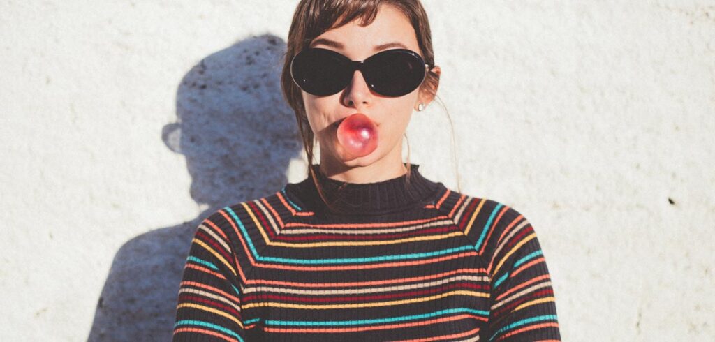 Eine Frau mit schwarzer Sonnenbrille macht eine Blase mit einem Kaugummi. (c) AdobeStock