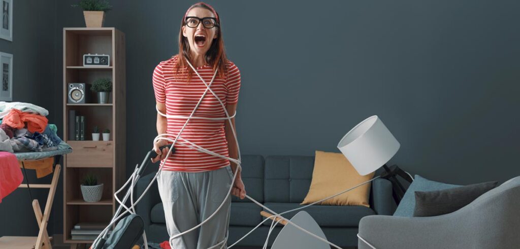 Eine Frau schreiend in einem unaufgeräumten Wohnzimmer mit dem Staubsaugerkabel gefesselt. (c) AdobeStock