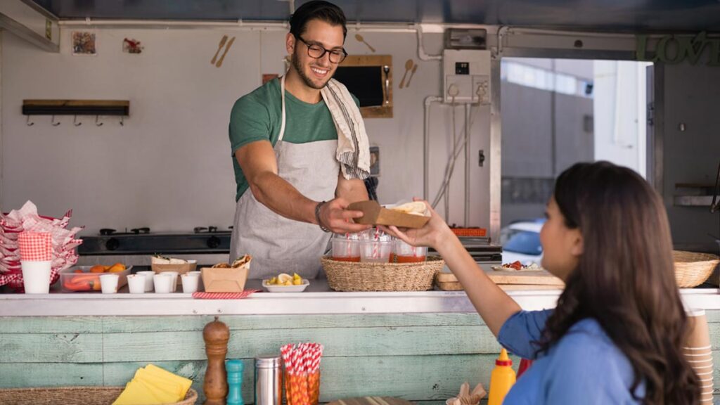 Ein Würstelbudenbetreiber gibt einer Kundin einen Wrap. (c) AdobeStock