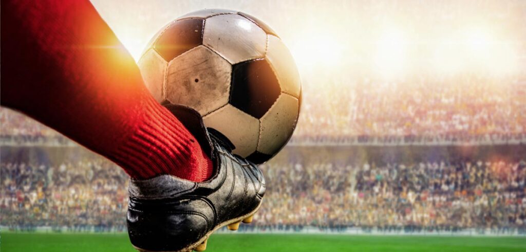 Close-Up eines Fußes mit Fußballschuhen, der einen Ball tritt; dahinter ein voll besetztes Stadion. (c) AdobeStock