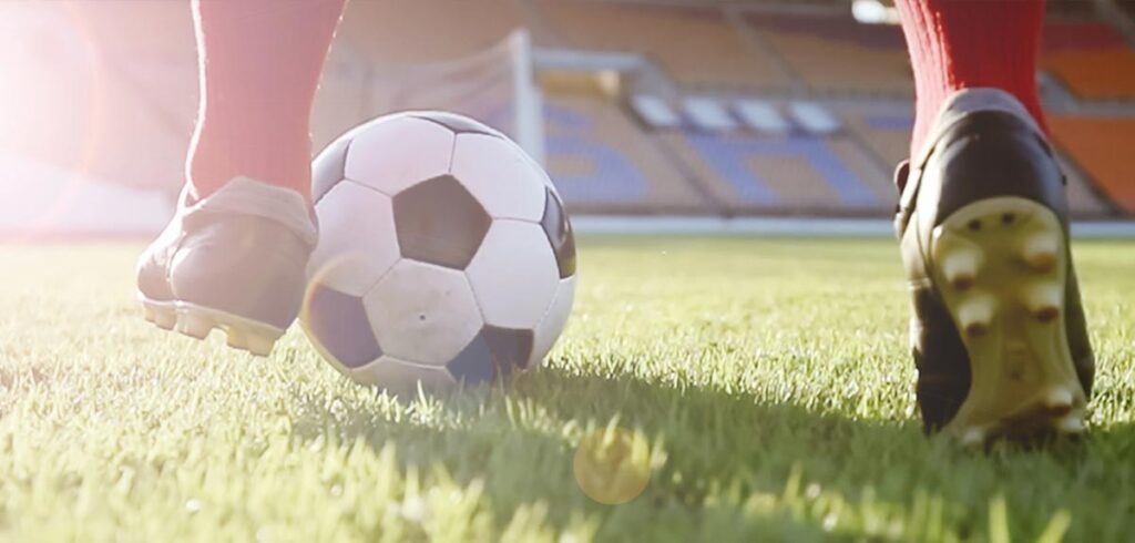 Die Beine eines Fußballers, der mit dem Ball auf einem Spielfeld rennt. (c) AdobeStock