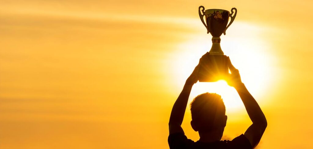 Silhouette eines Mannes, der einen Pokal in der Abendsonne in die Höhe stemmt. (c) AdobeStock