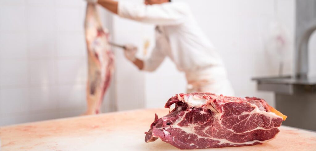 Ein Stück Fleisch, dahinter ein Fleischhauer bei der Arbeit. (c) AdobeStock