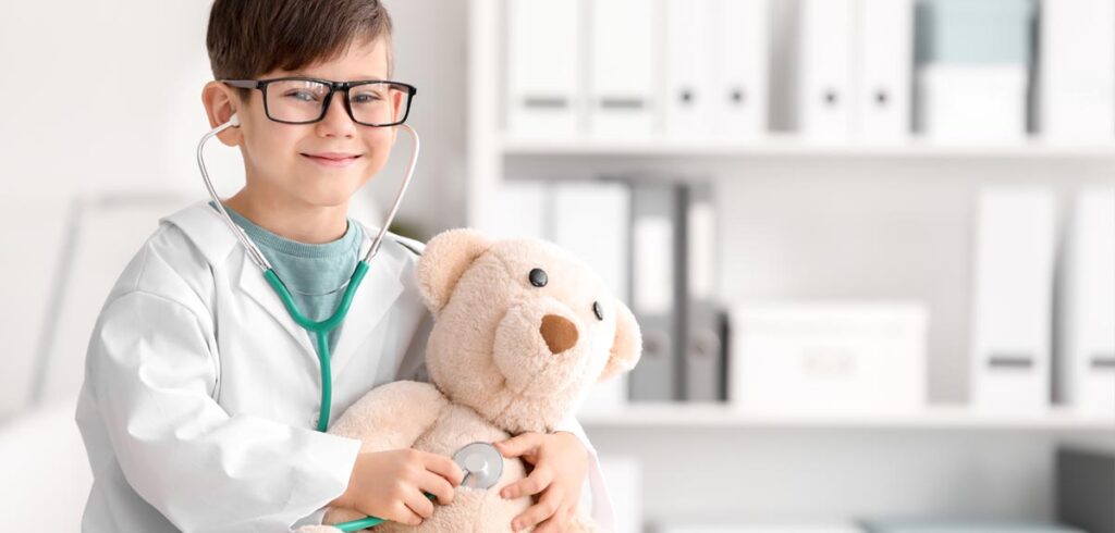 Ein Junge als Arzt mit Stethoskop, das er auf einen Teddybären hält. (c) AdobeStock