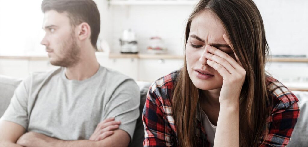 Ein Paar auf einer Couch; er mit verschränkten Händen in die eine Richtung schauend, sie genervt mit einer Hand vor den Augen in die andere. (c) AdobeStock