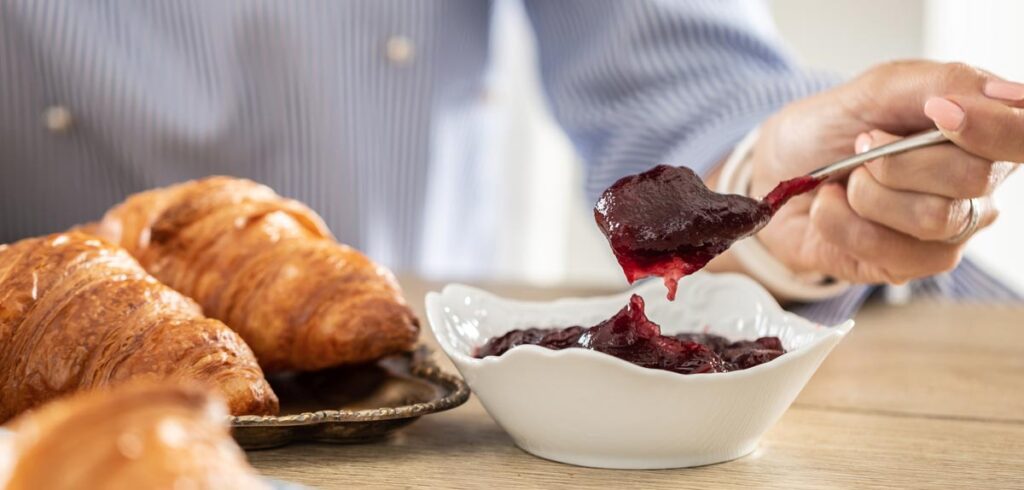 Die Hand einer Frau mit einem Löffel voller Marmelade, daneben Croissants. (c) AdobeStock