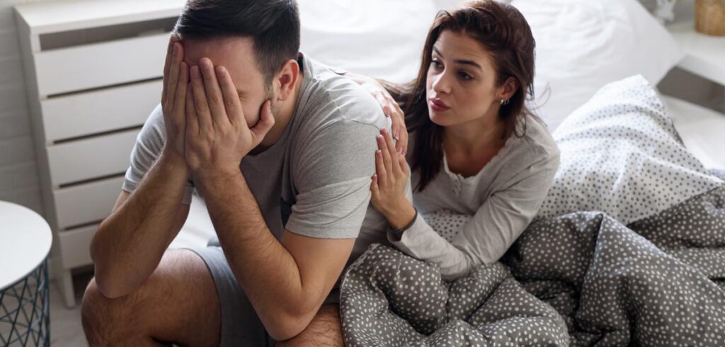 Ein Paar in einem Bett; er sitzt mit dem Kopf in seinen Händen; sie tröstet ihn. (c) AdobeStock