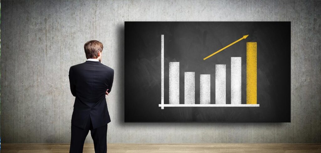 Ein Geschäftsmann steht mit verschränkten Händen vor einem Diagramm. (c) AdobeStock