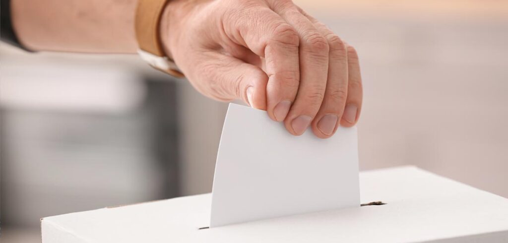 Die Hand eines Mannes, der einen Stimmzettel in eine Box wirft. (c) AdobeStock