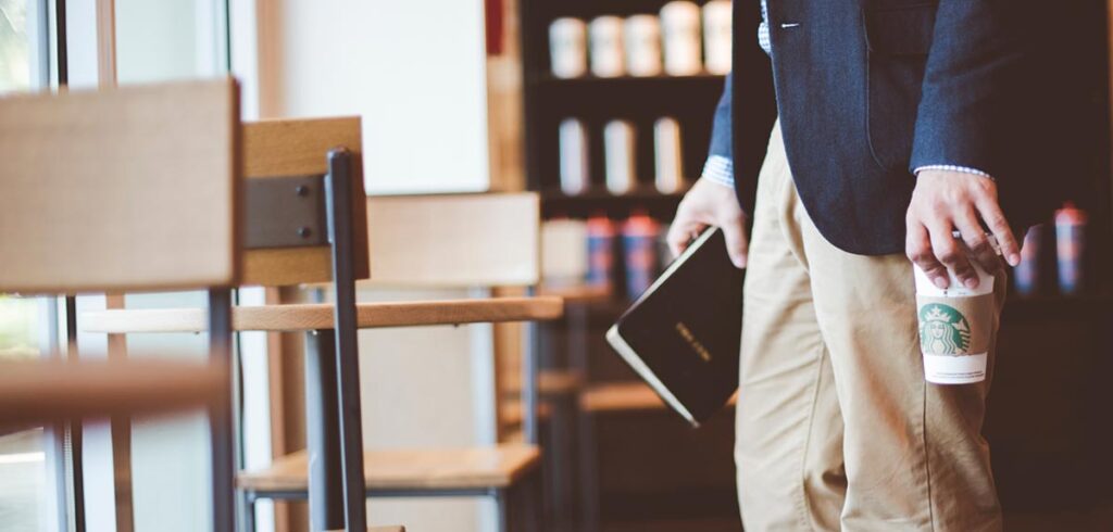 Ein Mann mit einem Starbucks-Becher in der einen und einem Notizbuch in der anderen Hand geht zu einem Tisch. (c) Pixabay.com