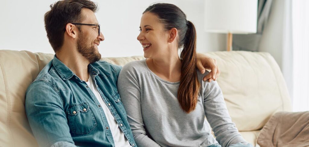 Ein Paar, das auf einer Couch sitzend sich gegenseitig anlacht und miteinander redet. (c) AdobeStock