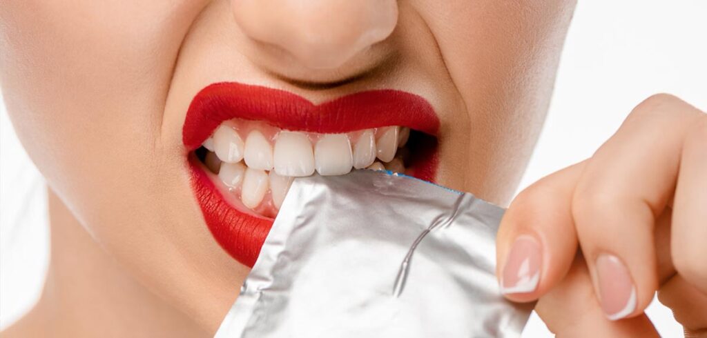 Der Mund einer Frau mit roten Lippen, die ein silbernes Kondom aufreißt. (c) AdobeStock