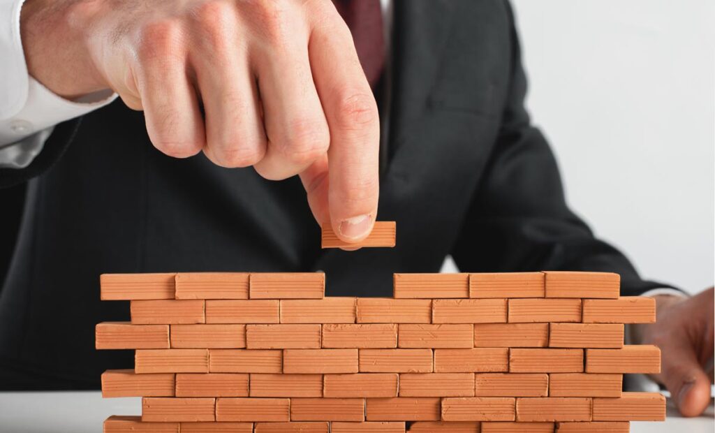 Die Hand eines Mannes, der mit Miniaturziegeln eine Mauer baut, Stichwort Koalition.
(c) AdobeStock