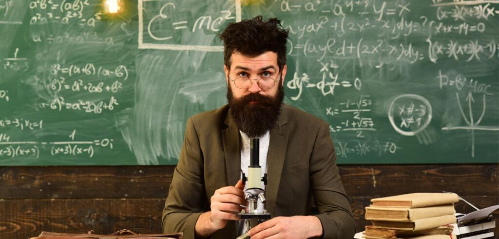 Ein Wissenschaftler mit Vollbart und Brille sitzt vor einer mit Formeln vollgeschriebenen Tafel. (c) AdobeStock