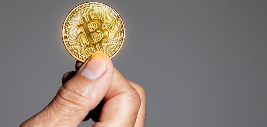 Die Hand eines Mannes, der einen goldenen Bitcoin hält. (c) AdobeStock