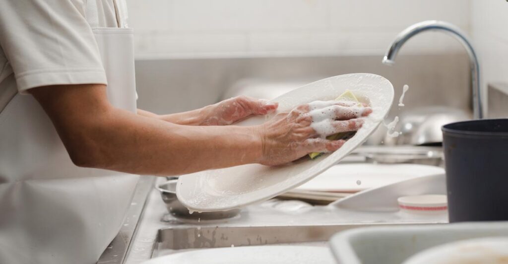 Die Hände eines Mannes, der einen Teller abwäscht, Stichwort Tellerwäscher.
(c) AdobeStock