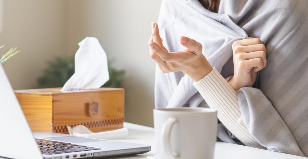 Nahaufnahme einer Frau, die am Schreibtisch vor einem Laptop sitzt, aber nur von der Brust bis zur Hand zu sehen ist. Sie ist in eine hellgraue Decke eingewickelt und trägt einen cremefarbenen Rollkragenpullover. Ihre rechte Hand ist leicht angehoben und die Handfläche zeigt nach oben. Links auf dem Tisch stehen eine Tasse und eine Holzbox mit Taschentüchern, Stichwort Regression.
(c) AdobeStock