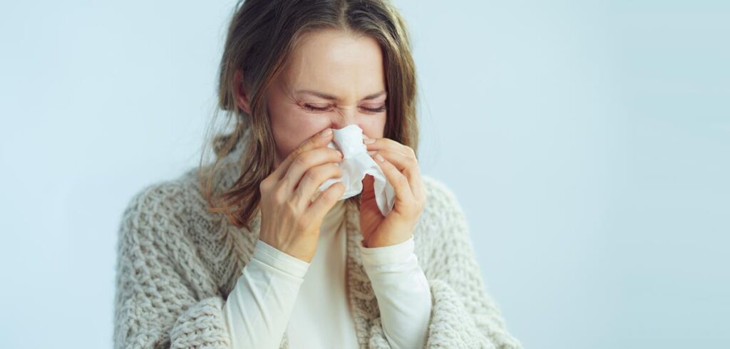 Eine Frau mit mittlerem, welligem Haar und geschlossenen Augen hält ein weißes Taschentuch an ihre Nase, während sie niesen muss. Sie trägt einen hellen, dicken, gestrickten Cardigan über einem weißen Rollkragenpullover vor einem hellblauen Hintergrund. (c) AdobeStock